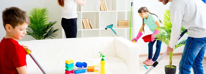 Twee volwassenen en twee kinderen maken schoon rond een witte bank waar diverse gekleurde schoonmaakspullen op liggen 