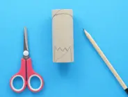 Dinosauruskop is getekend op een lege wc-rol met een schaar en een potlood aan weerskanten.