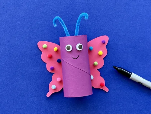 Een versierde vlinder van wc-rol met plakoogjes en een mondje getekend met de zwarte viltstift die ernaast ligt.
