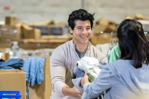 Een vrouw overhandigt een stapel kleren aan een man in een magazijn vol dozen met kleren.