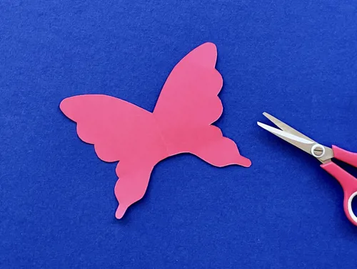 Uit roze karton geknipte vlinder met een schaar ernaast op een blauwe achtergrond.