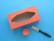 Oranje geverfde tissuedoos op een blauwe achtergrond met verf en kwast ernaast.