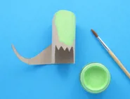 Deels groen geverfde kartonnen dinosauruskop met een staart eraan gelijmd en een verfpotje en penseel ernaast.