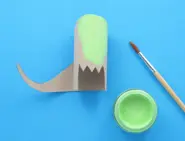 Deels groen geverfde kartonnen dinosauruskop met een staart eraan gelijmd en een verfpotje en penseel ernaast.