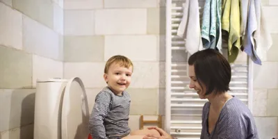 Een kind zit lachend op een wc-pot terwijl een vrouw met haar handen op zijn knieën lachend naar hem kijkt.