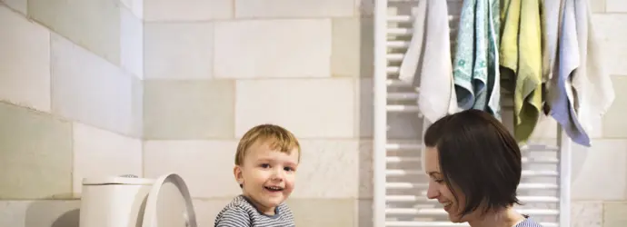 Een kind zit lachend op een wc-pot terwijl een vrouw met haar handen op zijn knieën lachend naar hem kijkt.