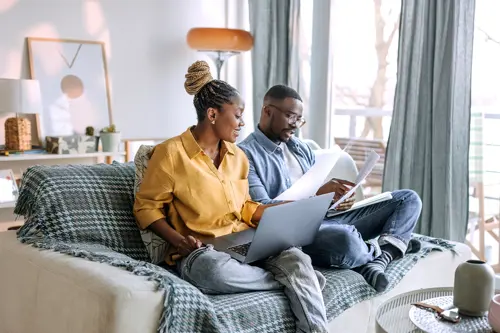 Een man en een vrouw zitten glimlachend op de bank terwijl ze papieren rekeningen doornemen en een laptop gebruiken.