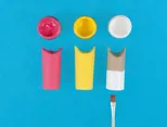 Een roze, een geel en een deels witgeverfd wc-rolletje met potjes verf en een penseel.