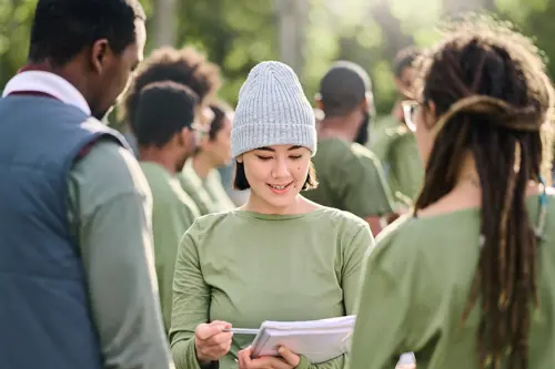 Een glimlachende vrouw, buiten, leest voor uit een document aan twee andere mensen.