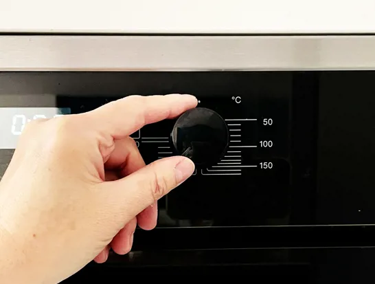 Een linkerhand draait een zwarte knop op een oven om de temperatuur in te stellen.