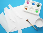 Een vel tekenpapier met repen gescheurd keukenpapier, verf op een palet, een kommetje lijm en een kwast.