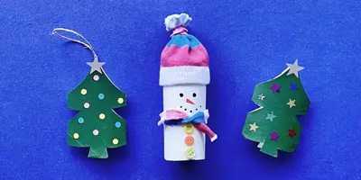 Twee versierde kerstboompjes en een sneeuwpop geknutseld van lege wc-rollen op een blauwe achtergrond.