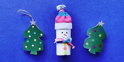 Twee versierde kerstboompjes en een sneeuwpop geknutseld van lege wc-rollen op een blauwe achtergrond.