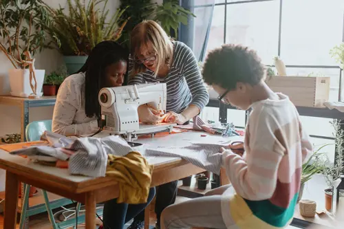 Twee vrouwen zitten bij een naaimachine, terwijl een jongetje aan dezelfde tafel in wat stof knipt.