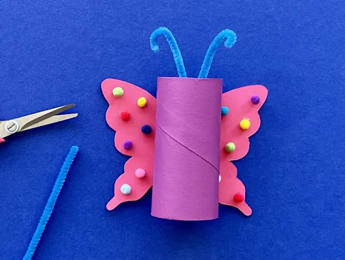 Een roze vlinder van wc-rol en karton met blauwe antennes van pijpenrager; een schaar en pijpenrager liggen ernaast.