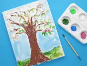 Een geverfde collage van keukenpapier en karton in de vorm van een boom met een verfpalet en een penseel ernaast.