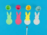 Vier kartonnen konijntjes die groen, roze, geel en blauw geverfd worden met potjes verf en een penseel.