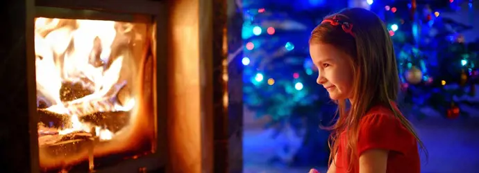 Meisje met lang haar kijkt naar een haardvuur achter een kachelruit met een verlichte kerstboom in de achtergrond.