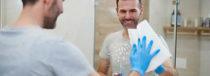 Glimlachende man met een blauwe handschoen aan veegt een spiegel schoon met een vel keukenpapier.