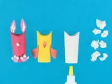 Drie geverfde wc-rollen waarop karton en propjes papier zijn geplakt om paasversiering te maken in de vorm van diertjes.