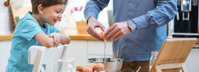 Een man en een kind breken vrolijk eieren in een mengkom.