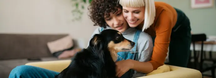 Twee vrouwen en een hond knuffelen op een mosterdgele bank.