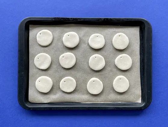 Twaalf uit zoutdeeg gesneden rondjes met een gaatje erin geprikt liggen op een met bakpapier bekleede bakplaat.